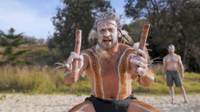 Gumbaynggirr man dressed for ceremony, holding clapsticks, facing camera