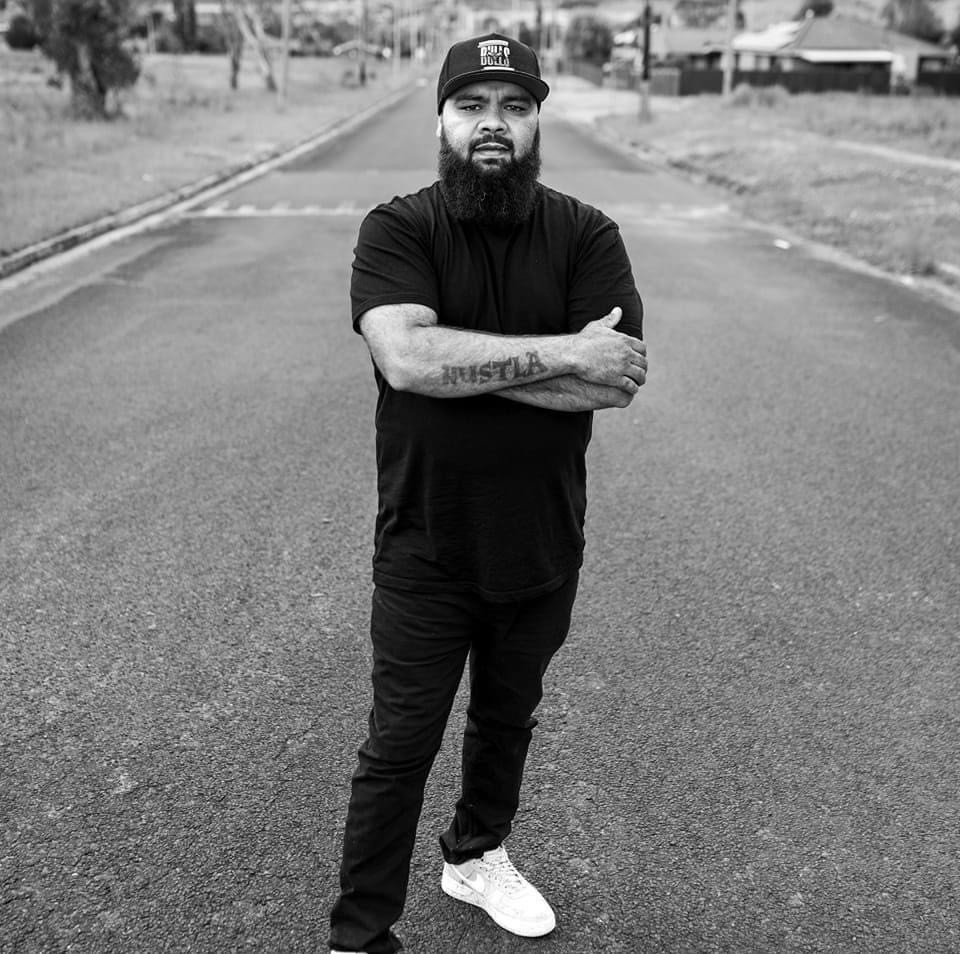 An aboriginal man dressed in a black baseball cap, a black t shirt and black pants stands in the middle of a road. He is looking straight at the camera and has his arms folded. A tattoo on his right forearm reads 'Hustla'