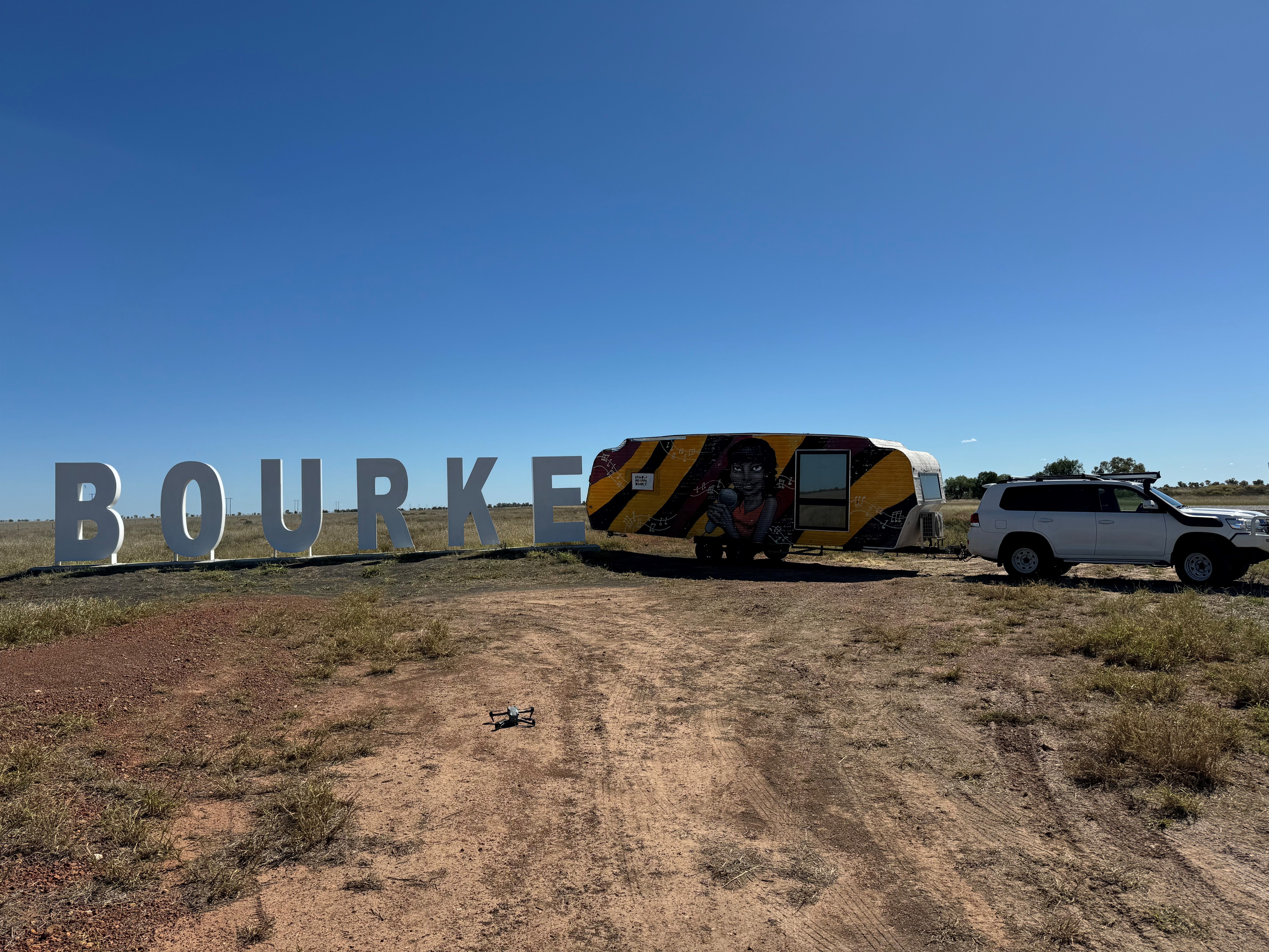 Large white letters spelling out 'Bourke' stand in a flat empty outdoor space. At the end of the sign is a four-wheel-drive with a caravan attached. The caravan is painted in yellow and black with an image of a person holding a microphone in the middle.