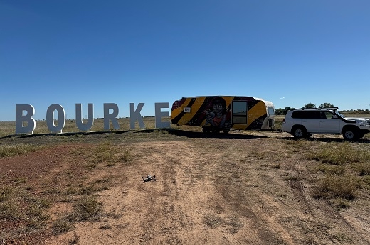 Large white letters spelling out 'Bourke' stand in a flat empty outdoor space. At the end of the sign is a four-wheel-drive with a caravan attached. The caravan is painted in yellow and black with an image of a person holding a microphone in the middle.