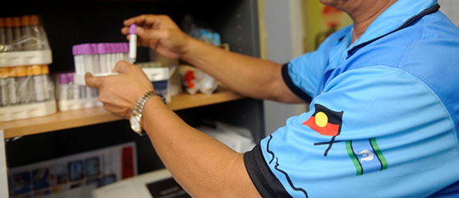 Pathology services — a person holding samples of bodily fluids. The person is shown close-up of their neck to elbows. Their shirt is printed with Aboriginal and Torres Strait Islander flags 