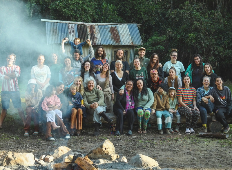 A large group of people poses for a photo. The front row is sitting and the middle and back row are standing. Smoke from a bonfire partly covers the people on the left of the image