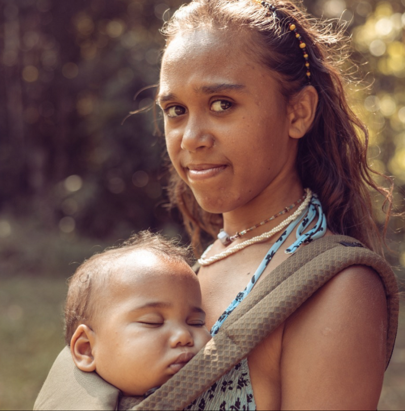 A close-up photo of a young woman. A baby is asleep in a baby carrier on her front