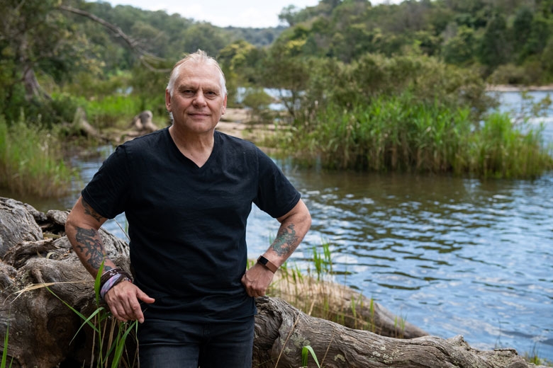 A man stands looking at the camera. Behind him is a river