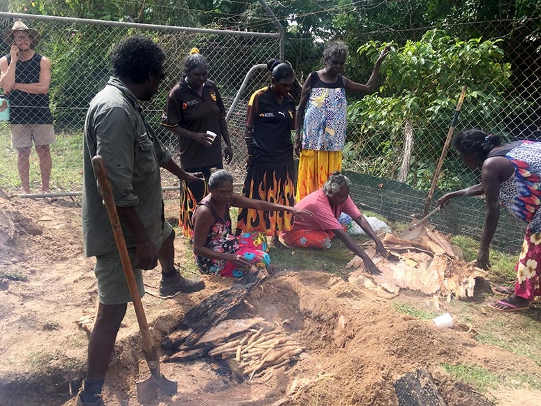 A group of people watch as women unwrap a paperbark parcel next to a fire pit