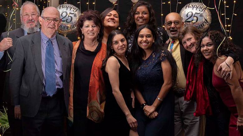 A group of well-dressed people pose. Behind them are balloons saying 25th
