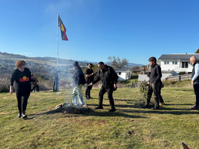 A group of people moving around a fire. An Aboriginal flag flies in the background
