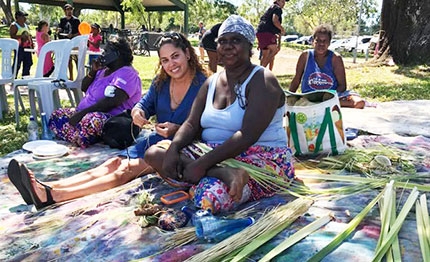 Two women sit on a mat surrounded by weaving materials