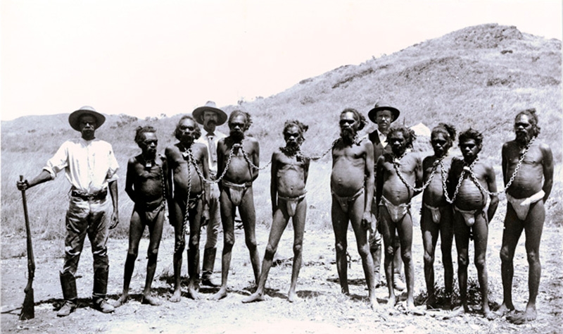 A group of Aboriginal men chained together by their necks. Several white men with rifles stand behind the group