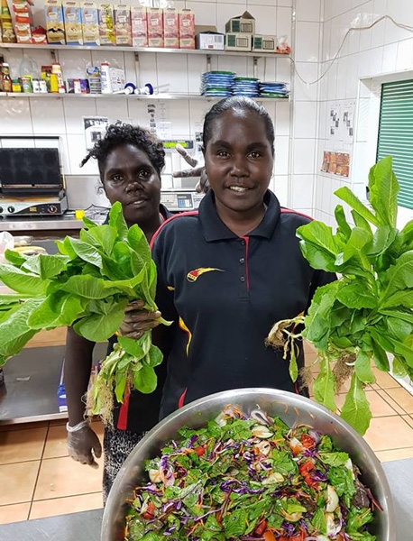 Two women are in a kitchen. One holds leafy vegetables. In front of them is a large bowl of salad