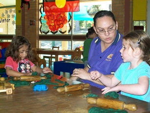 An adult sits with two children at a table. There is play dough and rolling pins on the table