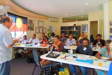 A group of adults sit at desks in a classroom. A man stands in front of them.