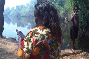 A woman stands by a river holding a walkie talkie. A boy stands near the water's edge
