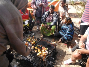 A person cooks food on a BBQ. Other people site on the ground nearby