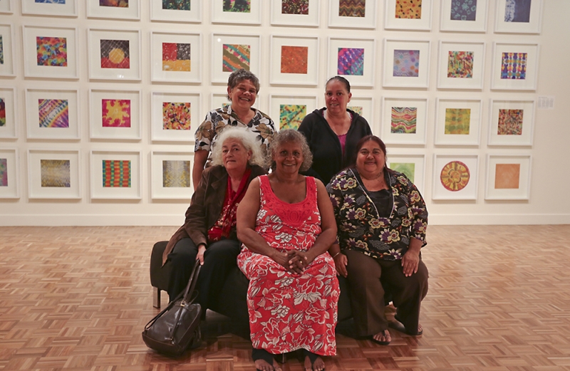 A group of women pose for the camera. Behind them is a large wall covered in artworks