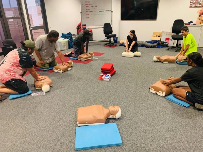 A group of people kneel on the floor in a room. In front of them are CPR dummies.