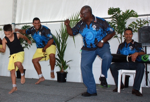 Men dancing on a stage. At the back a man is sitting and playing an instrument