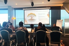 People sit at desks in 2 long rows, facing a screen that shows text and an image of a tree