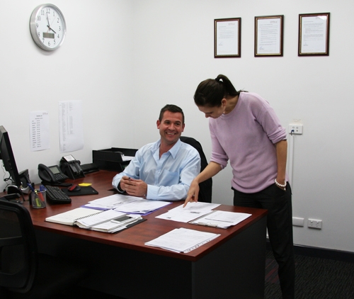 A man sits at a desk. A woman stands next to him