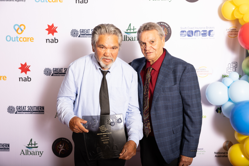 Two men pose in front of a wall covered in corporate logos. One man is holding an award