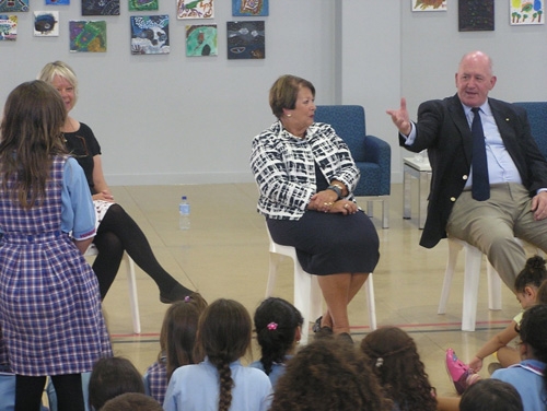 Children sitting on the floor looking at 3 people sitting on chairs facing them