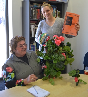 A woman sitting at a desk with flowers in front of her. Another woman stands beside her holding up a document