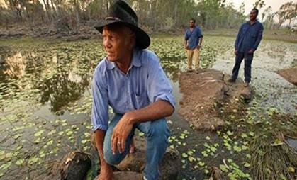A man wearing a hat crouches on a wetland. Two men are standing in the background
