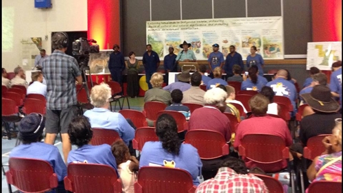 People stand at the front of a room behind a lectern. Other people sit in chairs facing them